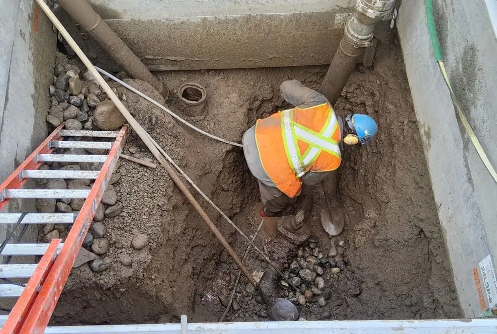 Débouchage Verviers et Liège : Un ouvrier utilise la méthode hydro-vac pour exposer en toute sécurité une vanne à guillotine enterrée à l'intérieur d'une tranchée tandis que les conduits électriques et de télécommunications restent sous tension au-dessus.